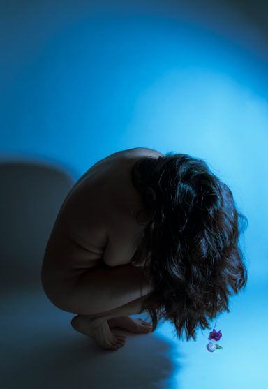 Nackte Person in zusammengesunkener Haltung mit blauer Beleuchtung und einer Blume.